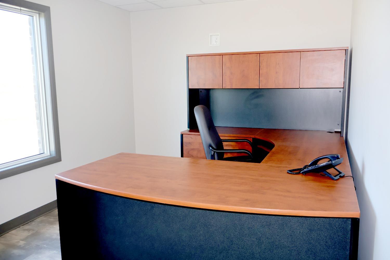 A single-person office with a large wooden desk, an ergonomic chair, and a telephone placed on the corner. The room has a window allowing natural light to enter.