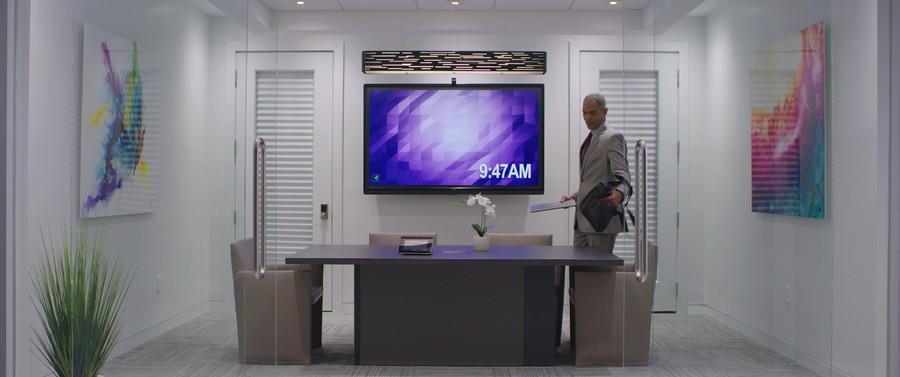 Boardroom featuring tabletop room control and large display screen with a man walking in and setting down his laptop