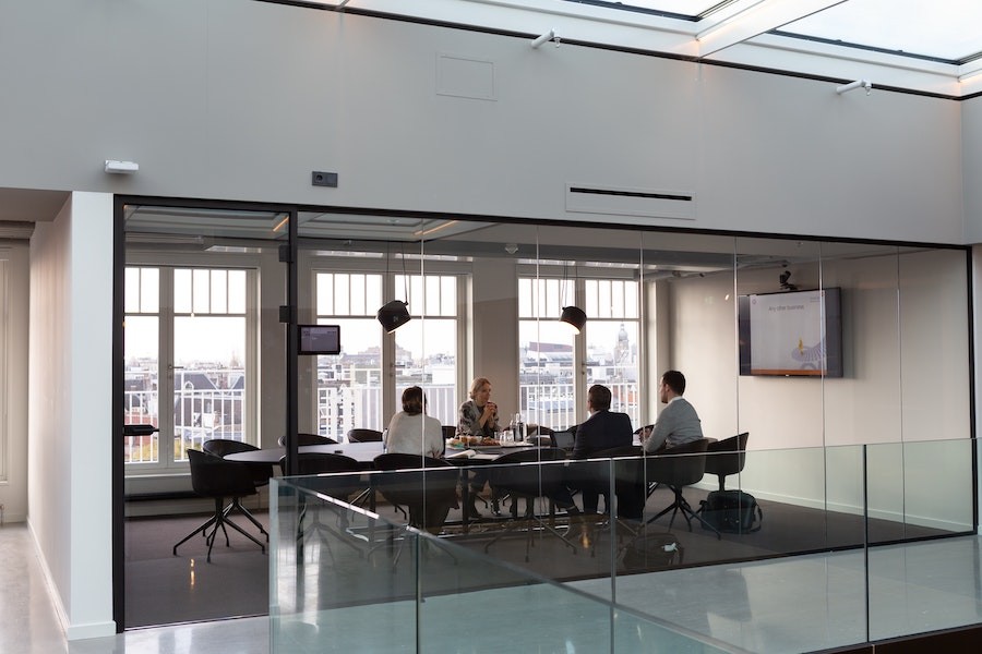 glass-enclosed conference room with four people sitting around the table looking at the display screen