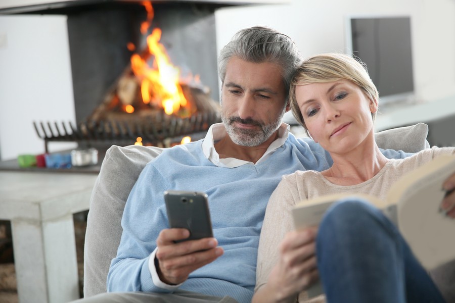 A couple sitting on the couch with a fireplace in the background, the man looking at his phone and the woman reading.