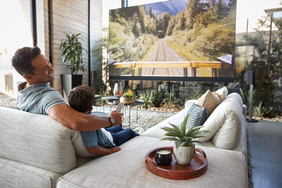 A father and son sitting on a couch watching a movie in a brightly lit living room.