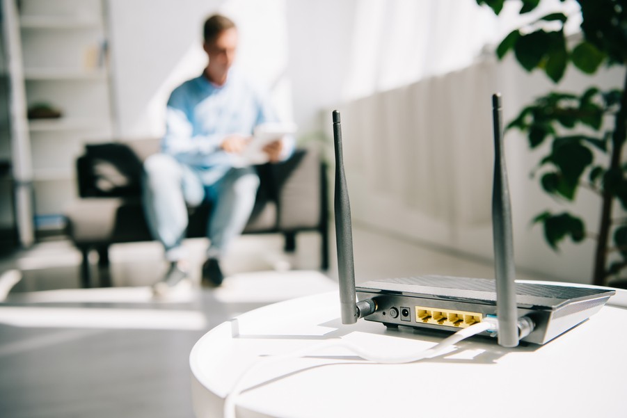 Image is of a home network router in the foreground with a person sitting in the background.