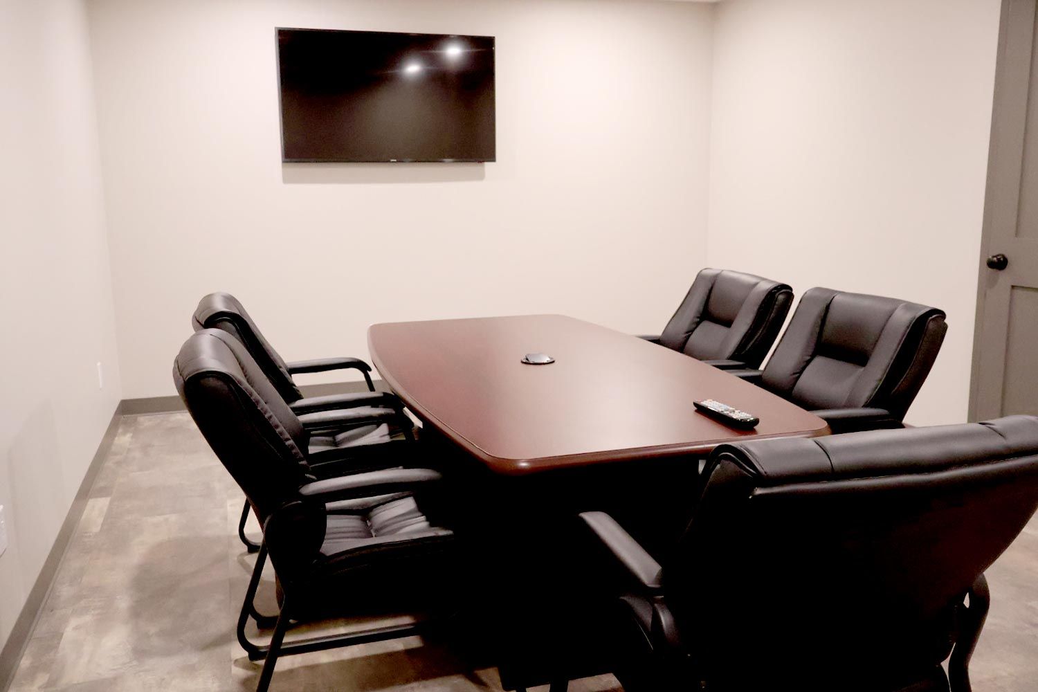 A compact meeting room with a rectangular wooden table surrounded by six black leather chairs. A wall-mounted TV screen is positioned at the front.