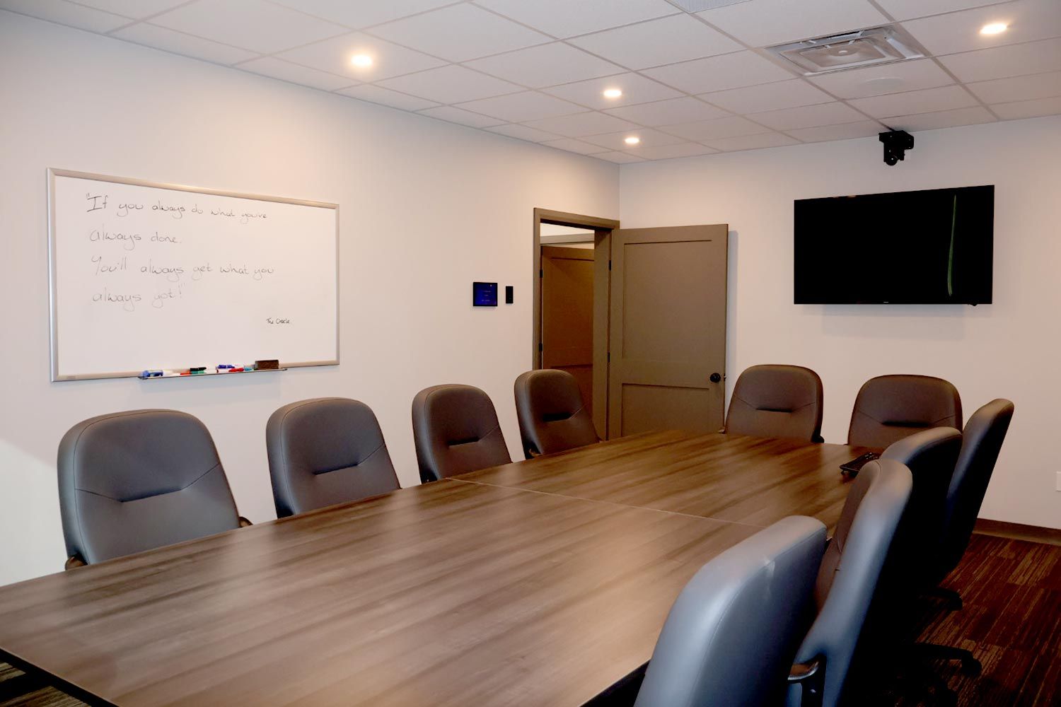 A spacious conference room with a long wooden table surrounded by multiple chairs. A large whiteboard with motivational writing hangs on the wall, and a TV is mounted in front.